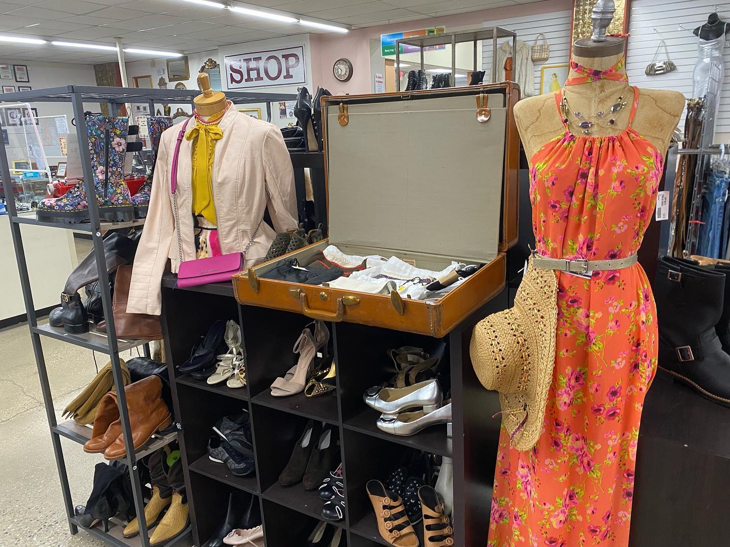 A display of the fashion and accessory Trunk Show at St. Vinny's Willy Street thrift store. There is a mannequin with a floral orange dress near an open trunk filled with colorful scarves. Next to the case there is a half-form mannequin wearing a light pink leather coat with a hot pink purse. Behind the half-form mannequin there is a tall rack of shoes.